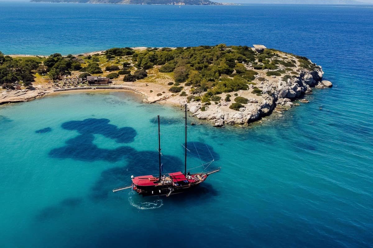 A two-masted schooner anchored off a pine-covered islet on an Aegina boat trip