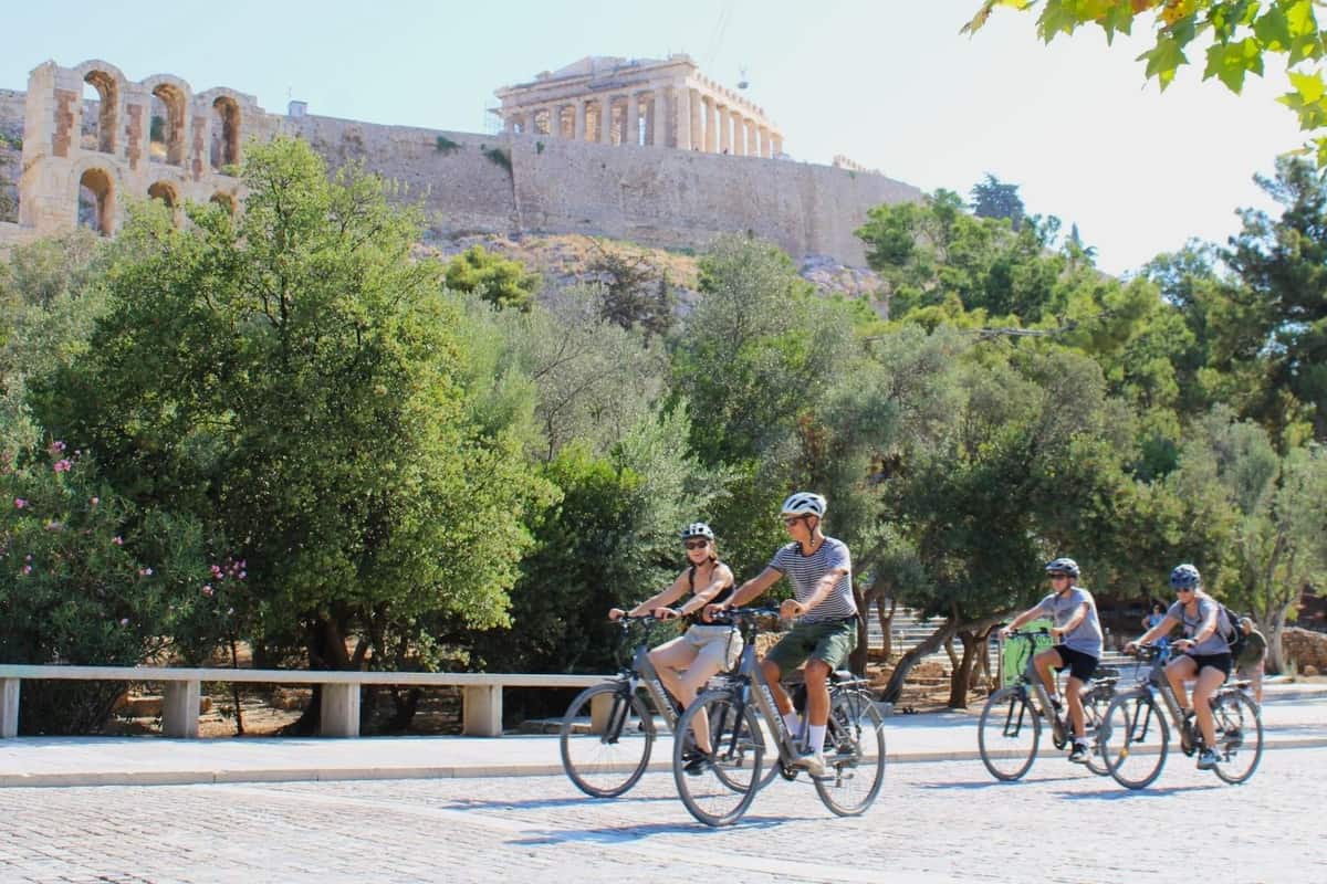 Riders on bikes pass along a paved street below the Acropolis and the Parthenon on an Athens bike tour