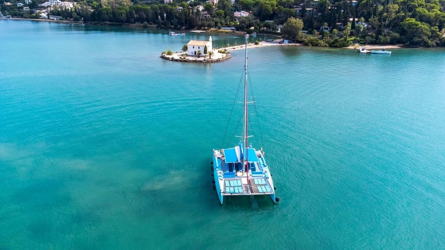 Catamaran on calm turquoise water near a small white chapel on a wooded headland, a Corfu sailing tour view
