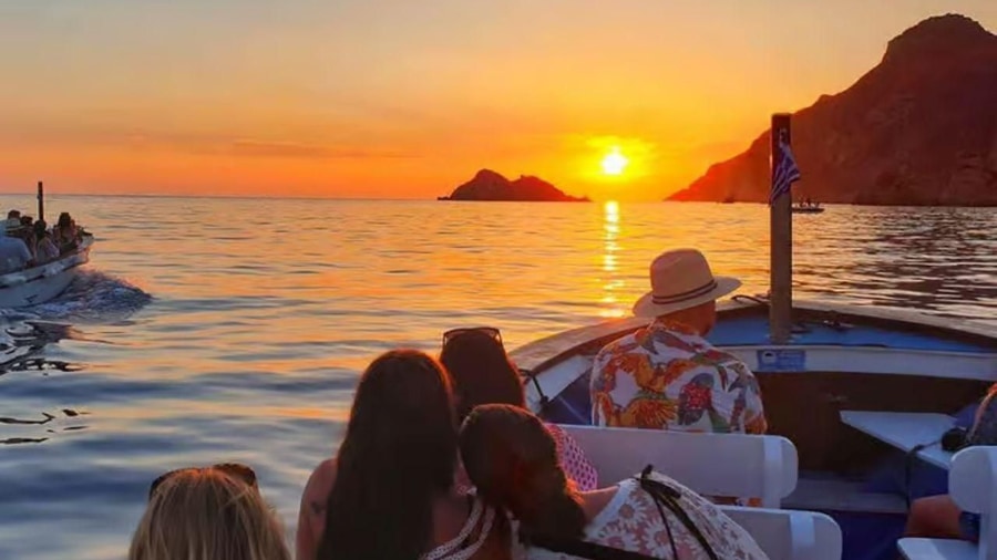Passengers on a wooden boat watch the sun set behind rocky islets and a headland on a Corfu sunset cruise