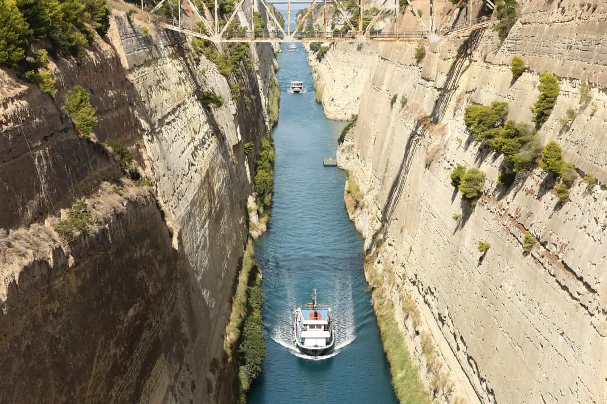 A passenger boat sails between the steep rock walls of the Corinth Canal under a steel bridge on a Corinth canal tour