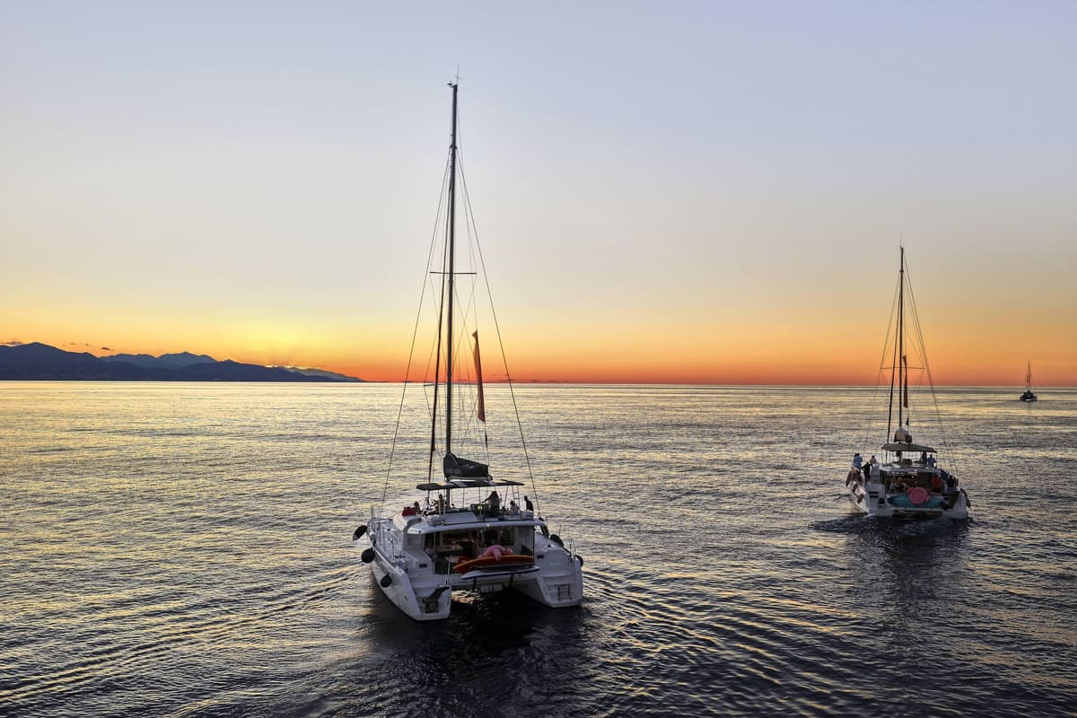 Two catamarans sail across calm water toward an orange sunset over Cretan mountains on a Crete sunset cruise