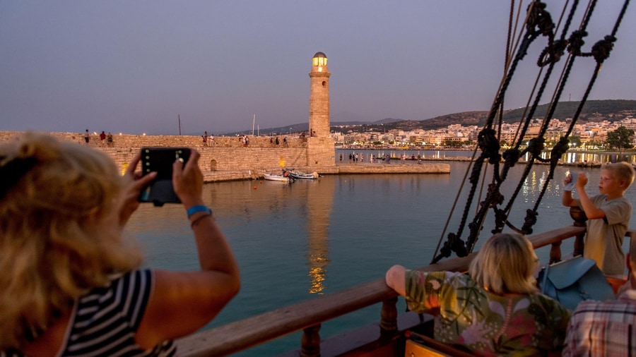 Passengers on a wooden ship photograph the lit Venetian lighthouse of Rethymno at dusk on a Crete sunset cruise