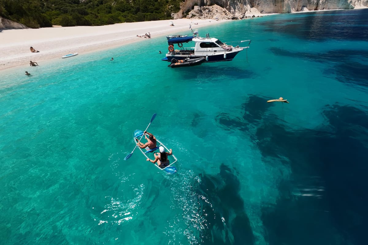 A motor boat and a clear kayak float in turquoise water off a white pebble beach on a Kefalonia boat tour