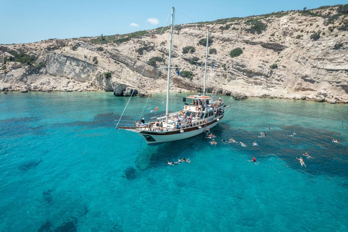 A wooden two-masted boat at anchor under white cliffs while swimmers float in turquoise water on a Kos boat trip