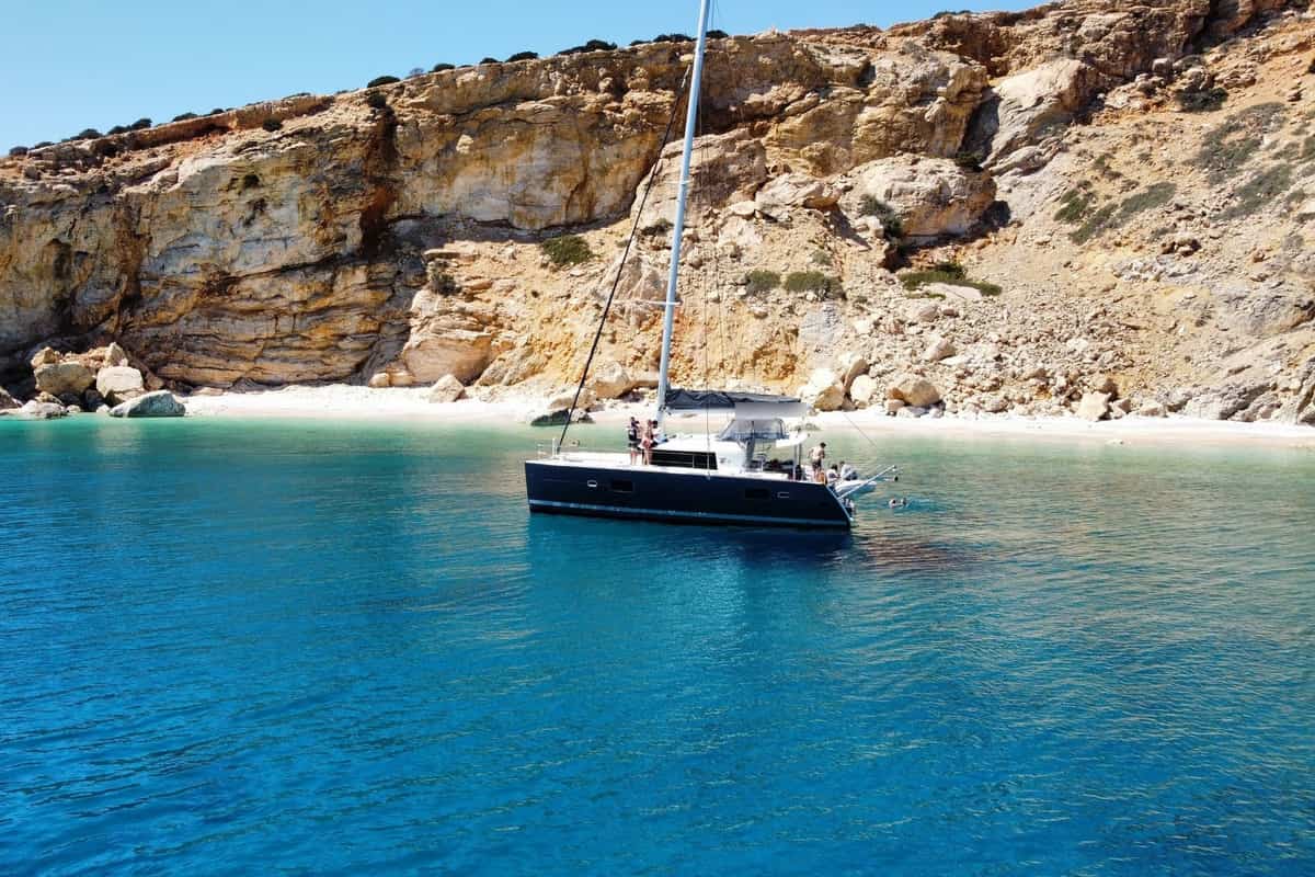 A catamaran on a Koufonisia cruise rests in a turquoise bay below golden cliffs and a white beach