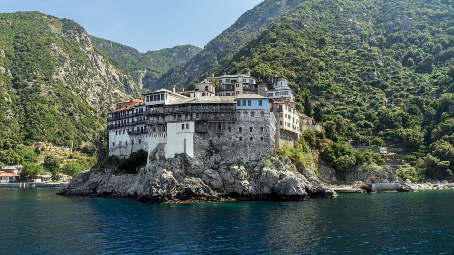 A tall stone monastery rises from a rocky point above the sea, below wooded hills, on a Mount Athos cruise