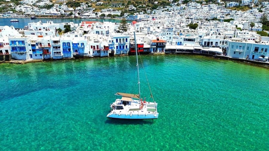 A catamaran floats in green-blue water off the white houses of Little Venice on a Mykonos catamaran cruise
