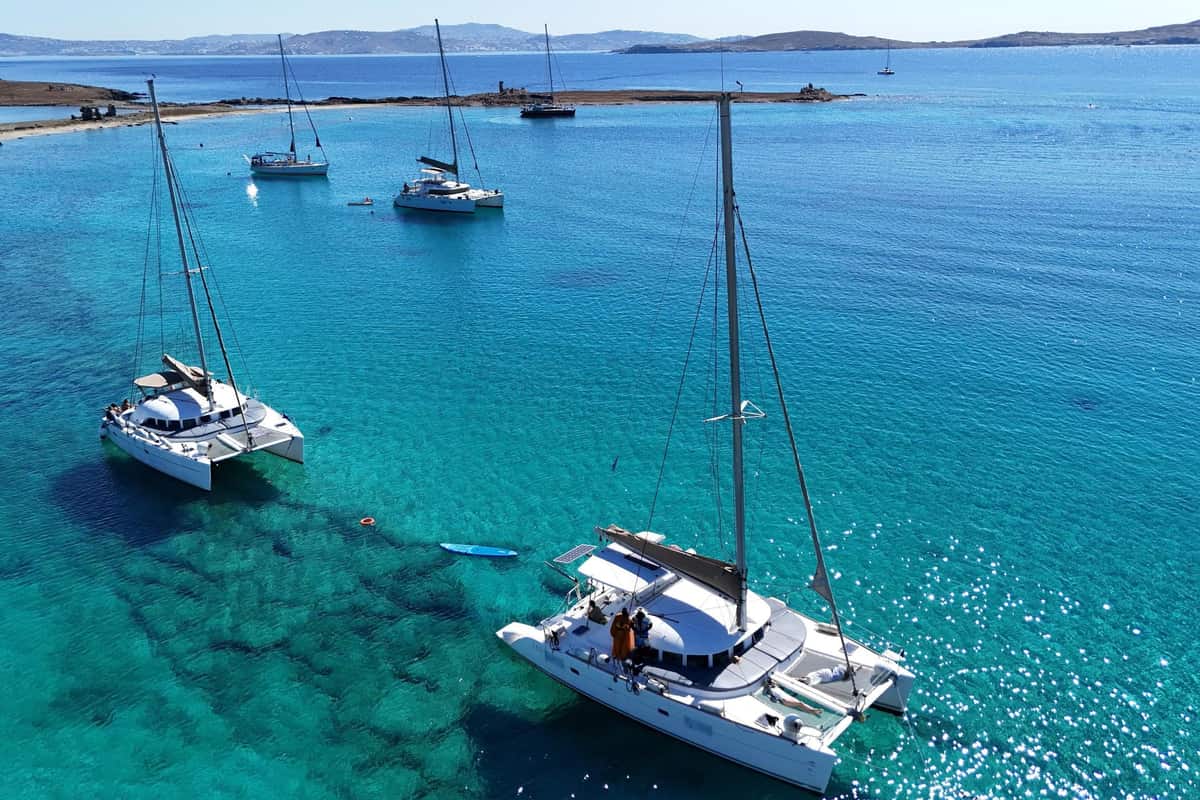 Sailing catamarans lie at anchor in a clear turquoise bay with a paddleboard nearby on a Mykonos catamaran cruise