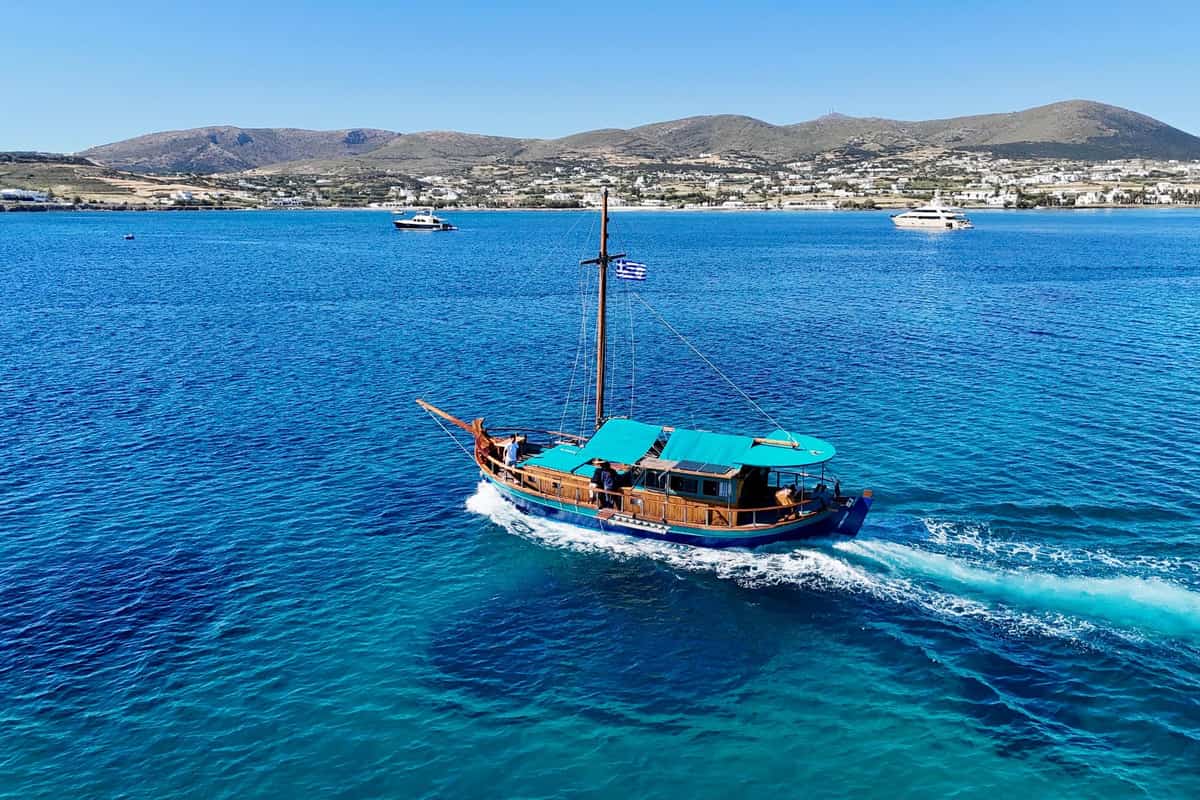 A wooden caique with a turquoise awning crosses deep blue Naoussa bay with the Paros coast behind on a Paros boat trip