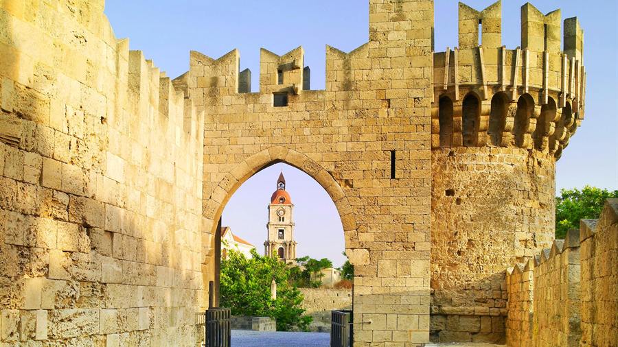A pointed gate in the medieval walls framing the clock tower of the old town, a first stop on a Rhodes shore excursion