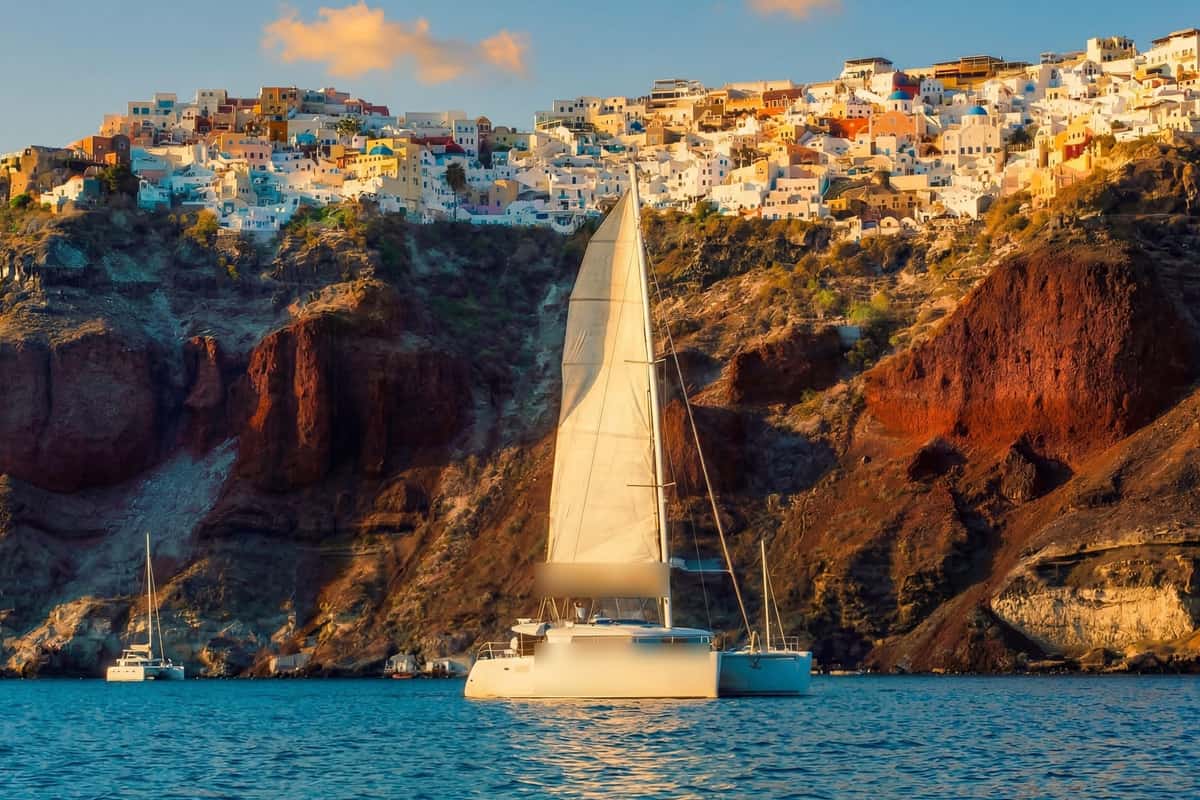A white sailing catamaran on a Santorini boat trip below the red cliffs and white houses of Oia