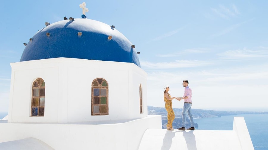 A couple holds hands on a white roof beside a blue church dome on a Santorini photo tour