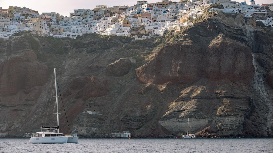 A catamaran sails below the red and grey cliffs of Oia on a Santorini sailing tour