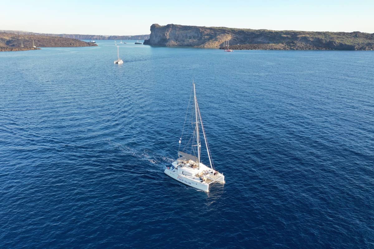 A sailing catamaran crosses the deep blue caldera with Thirasia behind on a Santorini sailing tour