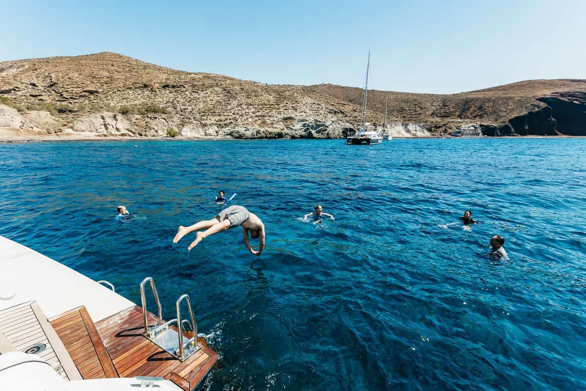 A swimmer dives off the stern of a catamaran into deep blue water off the coast while others swim on a Santorini snorkeling trip