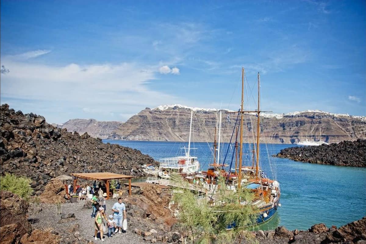 The black lava shore of a volcanic island with wooden boats and the caldera wall behind, seen on a Santorini volcano tour