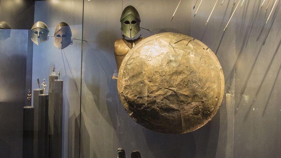 Bronze Corinthian helmets, a round bronze shield and spears in a museum case, the gear behind a Thermopylae tour story